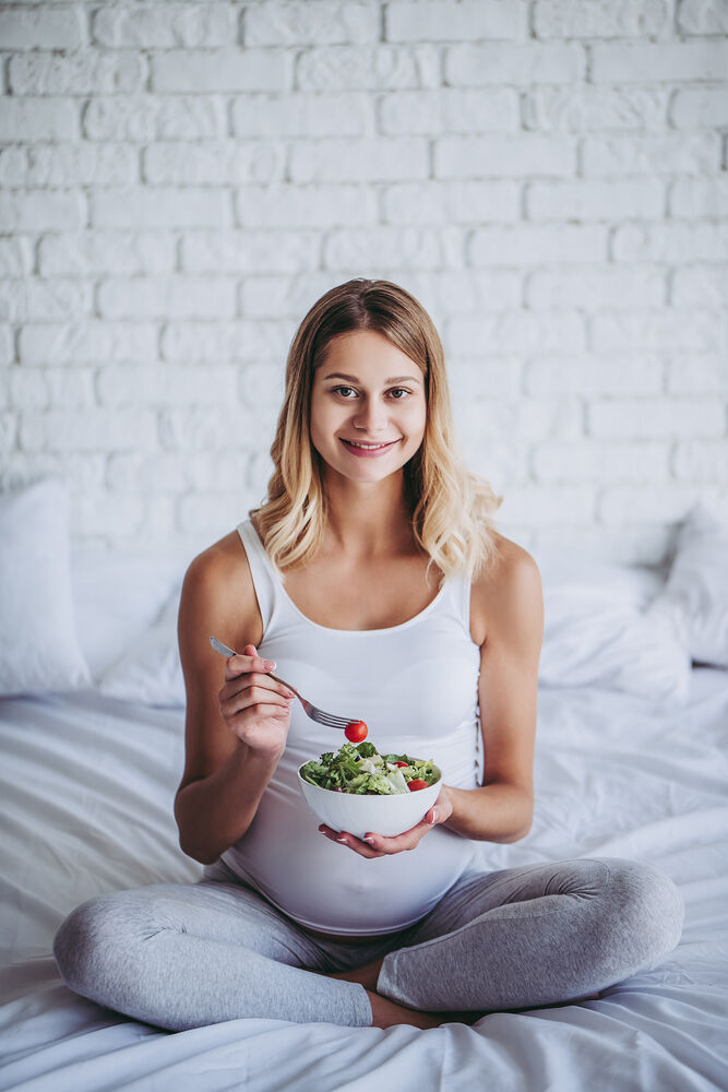 pregnant woman is sitting in bed and eating green salad.