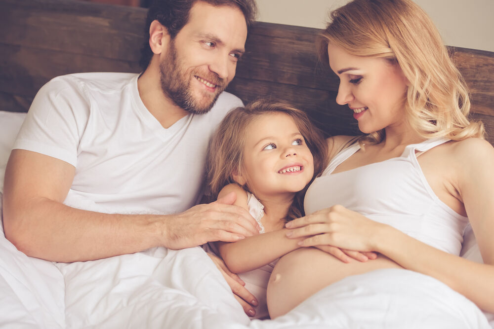  pregnant woman, her handsome husband and cute little daughter are talking and smiling while spending time together in bed