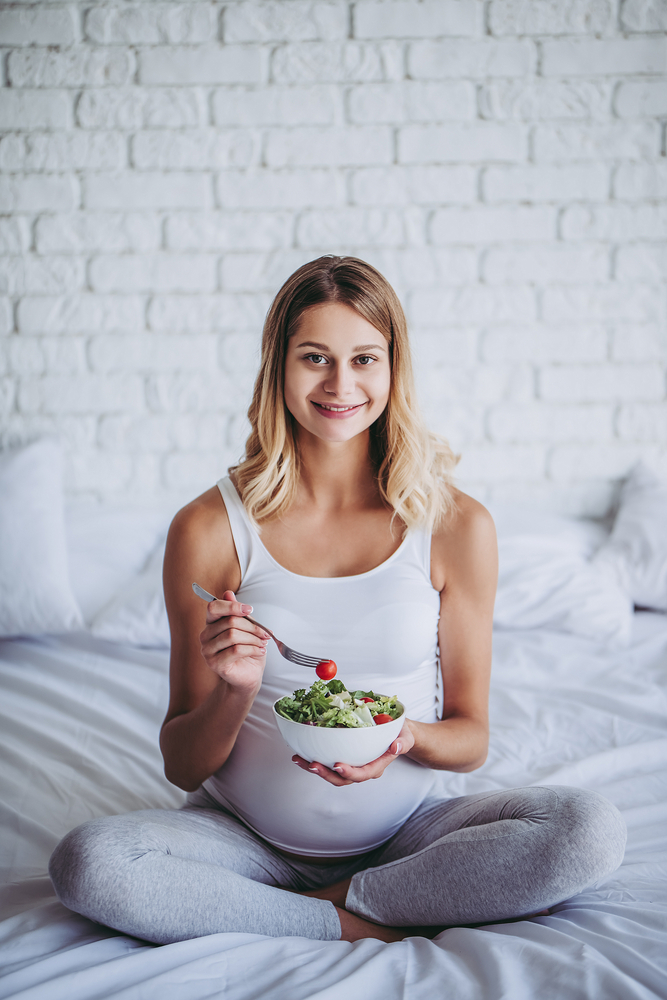 pregnant woman is sitting in bed and eating green salad. pregnant woman is sitting in bed and eating green salad.
