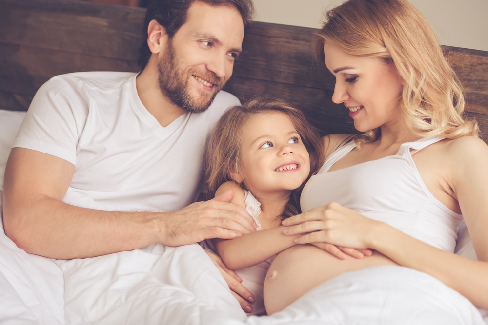 pregnant woman, her handsome husband and cute little daughter are talking and smiling while spending time together in bed  pregnant woman, her handsome husband and cute little daughter are talking and smiling while spending time together in bed