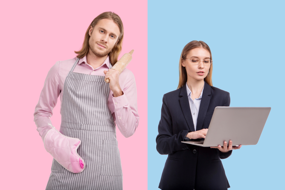 Destroying gender stereotypes. A man in an apron posing with a rolling pin, while his girlfriend is working on a laptop Destroying gender stereotypes. A man in an apron poses with a rolling pin, while his girlfriend works on a laptop